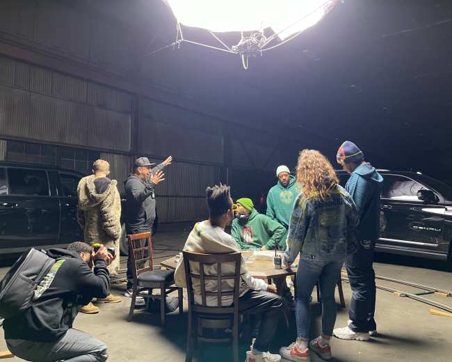 A group of people is gathered around a table inside a large, industrial space illuminated by overhead lighting, with vehicles parked nearby.