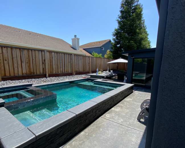 The image shows a backyard with a rectangular swimming pool and a spa area, surrounded by a wood fence and a concrete patio.