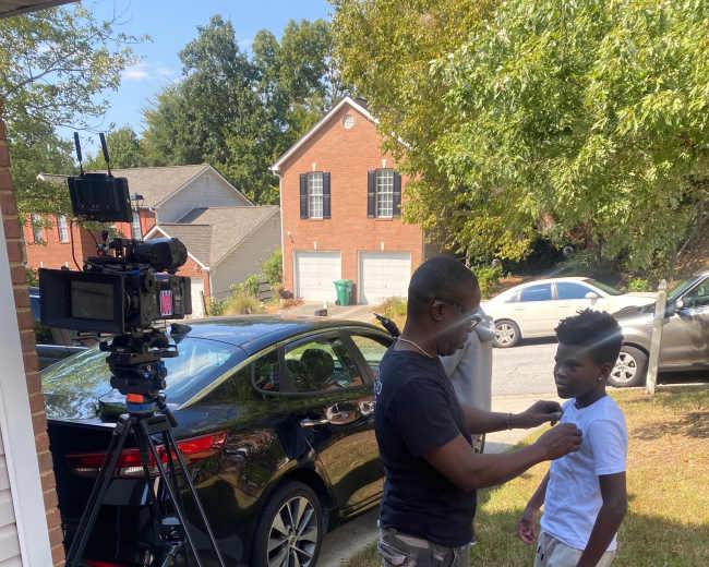 A person adjusts a microphone on a child while a video camera is set up nearby in a residential driveway.