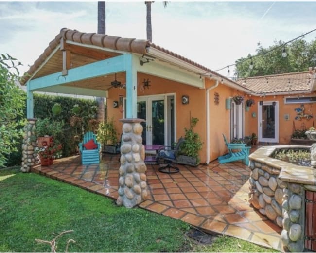The image shows a colorful orange house with a stone patio, surrounded by greenery and featuring an outdoor seating area with chairs.