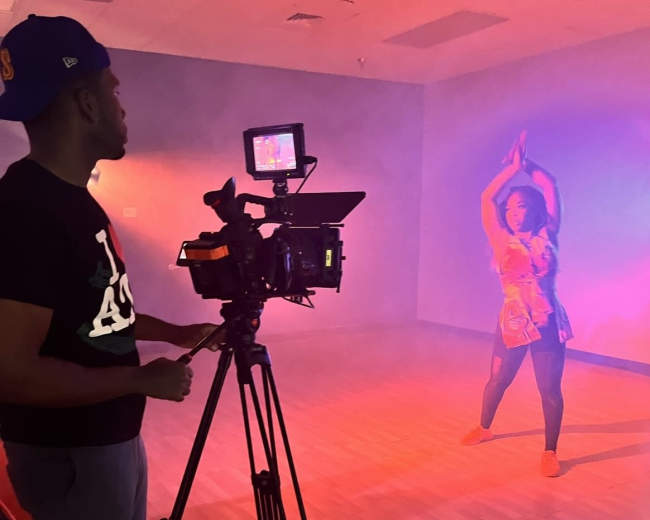 A person operates a camera while a dancer performs in a dimly lit studio filled with colorful lighting.