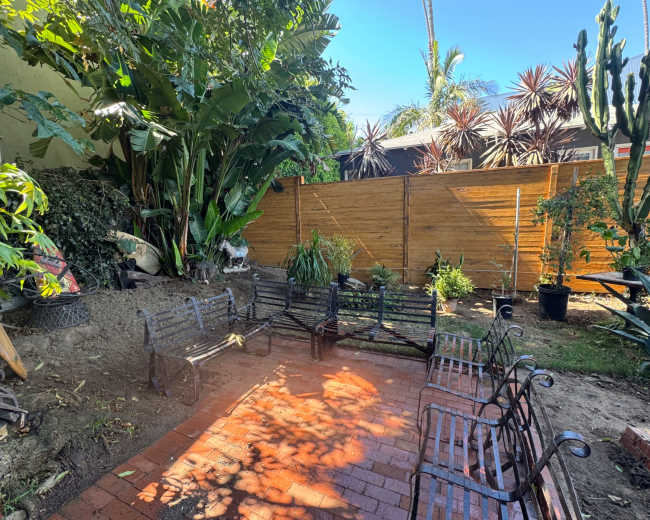 The image shows a small outdoor space with a brick patio surrounded by lush greenery and wooden fencing.