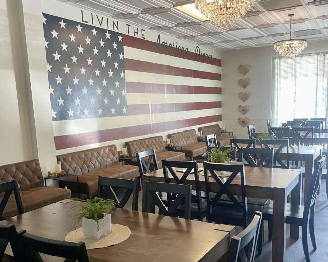 The image shows a restaurant interior featuring wooden tables, leather booths, and a large wall mural of the American flag.