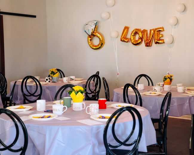 A decorated room features several round tables set with tablecloths, mugs, and floral centerpieces, with a balloon display spelling "LOVE" on the wall.
