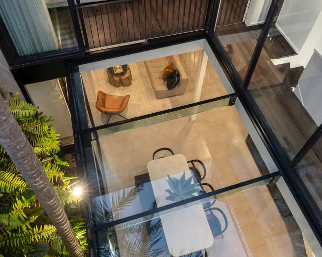 The image shows a modern multi-level interior space with a glass floor above a dining area, surrounded by indoor plants and characterized by sleek furniture and an open layout.