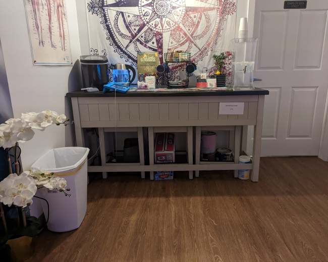 A beverage station with a counter, coffee machine, various drink supplies, and a trash bin, situated near a closed door and decorated with a large mandala.