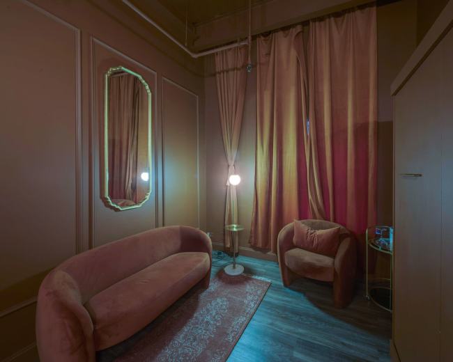 A small, dimly lit room featuring a curved sofa, an armchair, a mirror, a floor lamp, and curtains, all in warm tones.