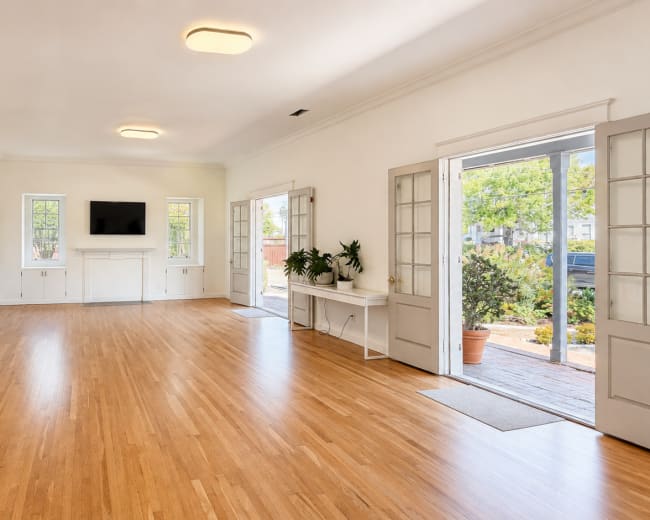 The image shows a spacious, empty room with hardwood floors, white walls, and large windows, leading to an outdoor area.