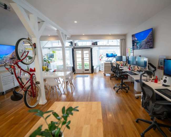 The image shows a modern office space with several desks, computers, and a bicycle hanging on the wall, illuminated by natural light through large windows.