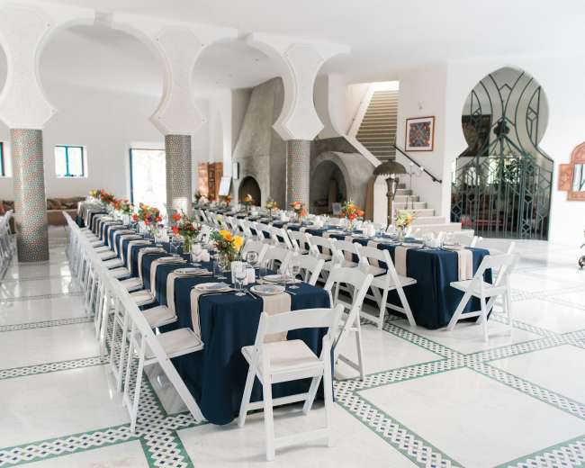 A spacious dining area features long tables set with navy blue tablecloths, white chairs, and floral centerpieces in a well-lit, modern room with arched columns.