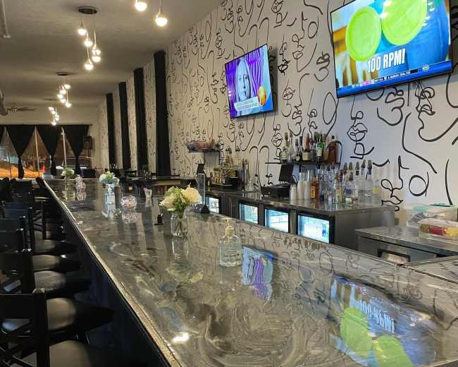 The image shows a modern bar with a marble countertop, featuring a variety of liquor bottles on shelves behind it, and two televisions mounted on the wall.