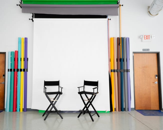 The image shows a photography studio setup featuring two black director's chairs, positioned in front of a large white backdrop, with colorful rolls of backdrop paper stacked beside the walls.