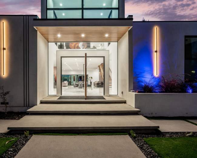 A modern home with a large glass entrance, flanked by bright lights and framed by landscaped pathways.