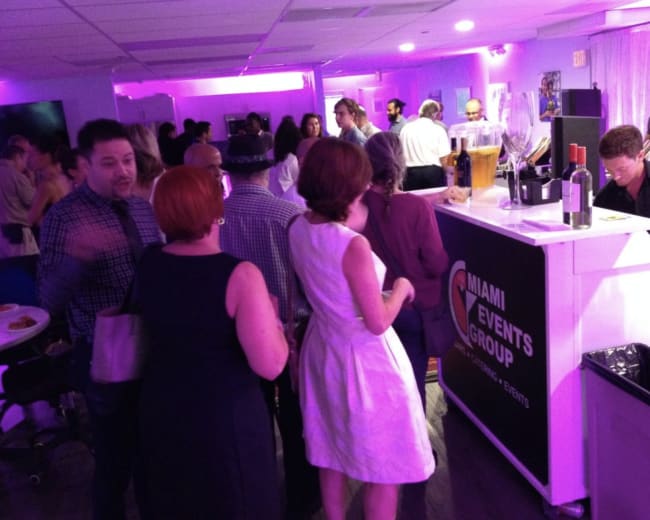 A crowd of people mingles at a brightly lit event space with purple lighting, gathered around a bar serving drinks.