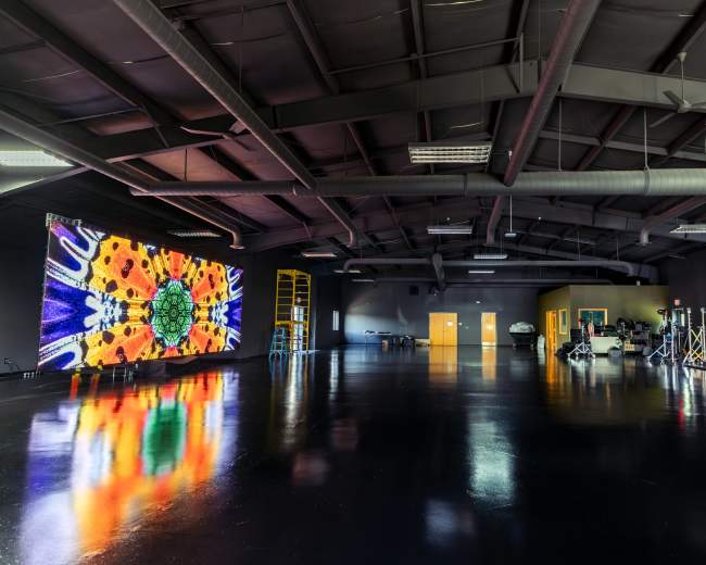 The image shows a spacious industrial room with shiny black floors, featuring a large colorful digital display on one wall and various equipment set up for audiovisual production.