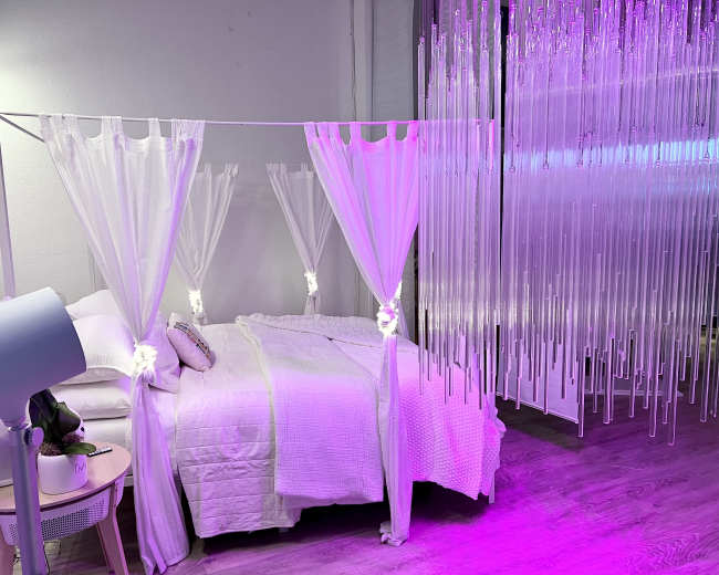 A bedroom features a bed with a white blanket and sheer curtains draped overhead, illuminated by soft purple lighting from hanging decor.