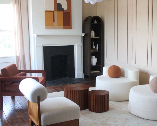 A modern living room features a cozy arrangement of sculptural chairs and circular tables around a simple fireplace, with a wall art piece in the background.
