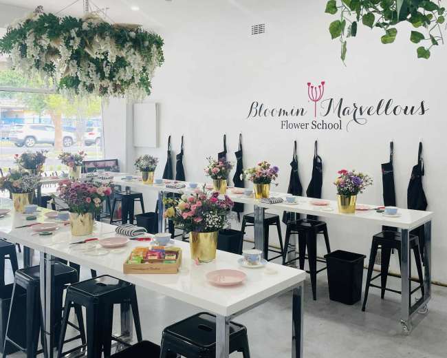 The image shows a modern flower school with white walls, several tables set with flower arrangements, and decor featuring hanging plants.