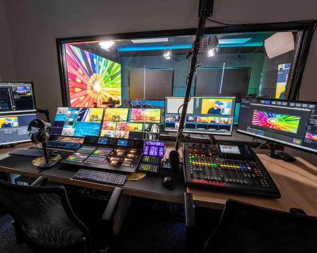 The image shows a broadcast studio control room filled with multiple computer monitors, a soundboard, and various control devices, all arranged on a large desk.
