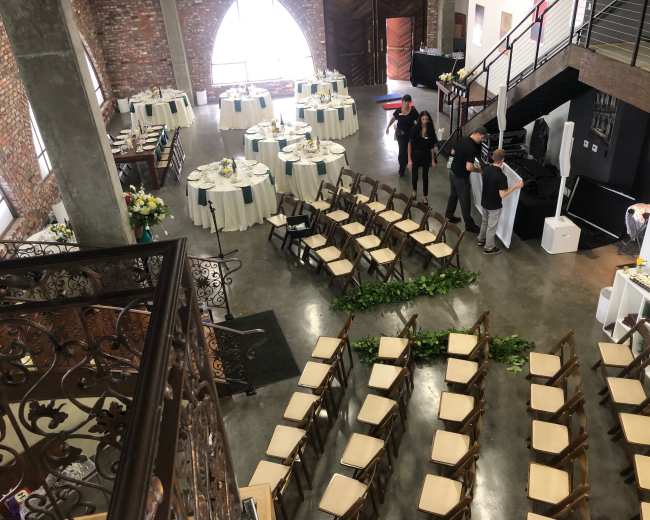 The image shows a multi-level event space with dining tables arranged for a reception and chairs set up for a ceremony beneath large arched windows.