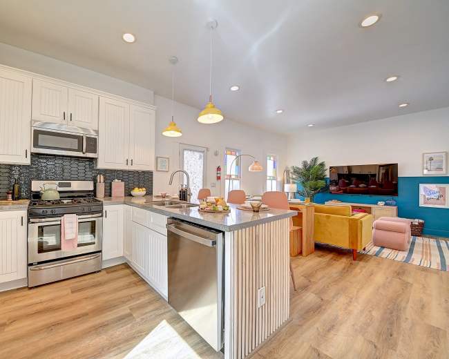 The image shows a modern kitchen with white cabinetry, stainless steel appliances, and a granite countertop, leading to a living area with colorful furniture and decorative art.