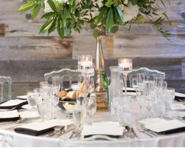 A round dining table is elegantly set with crystal glassware, rolled napkins, and a tall floral centerpiece, surrounded by clear chairs.