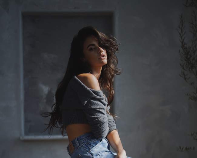 A woman with long hair sits sideways on a black stool, wearing a cropped sweater and high-waisted jeans, in a softly lit room.