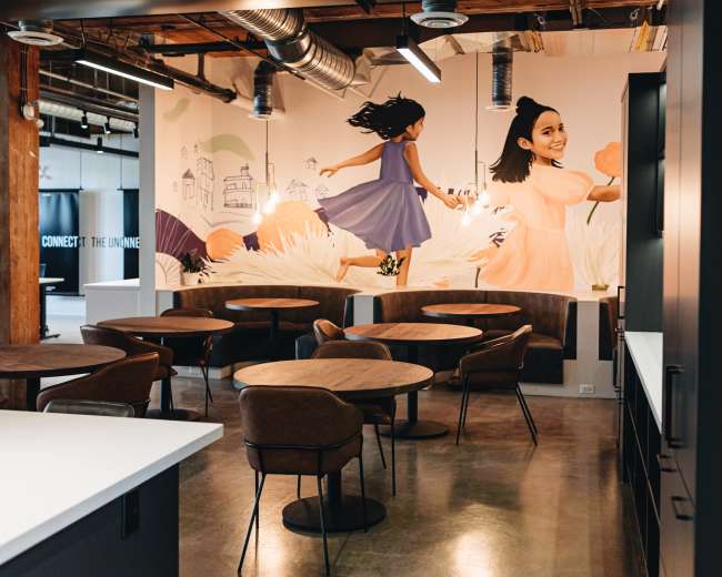 The image shows a modern office space with wooden tables and chairs, featuring a mural of a girl running on the wall behind.