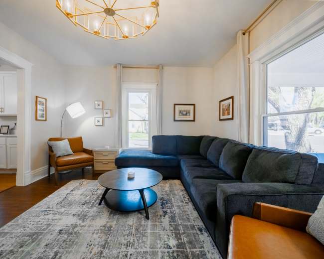 The living room features a large sectional sofa, a round coffee table, and a modern light fixture, with windows allowing natural light to enter.