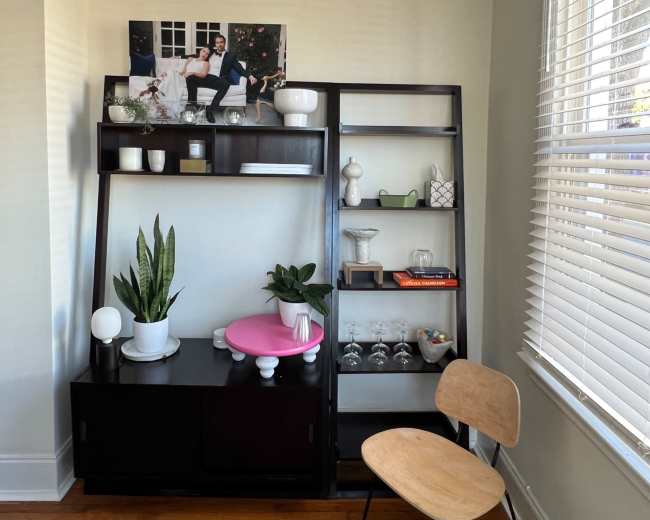 The image shows a black shelving unit with various decorative items, including plants and glassware, next to a wooden chair in a well-lit room.