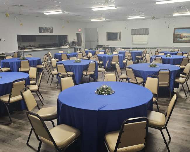 A large room is set up with multiple round tables covered in blue tablecloths, each adorned with a small floral centerpiece.