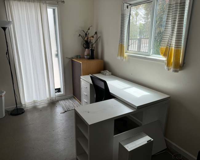 A simple office space with a white desk, a black chair, a cabinet, a lamp, and a window with sheer curtains.