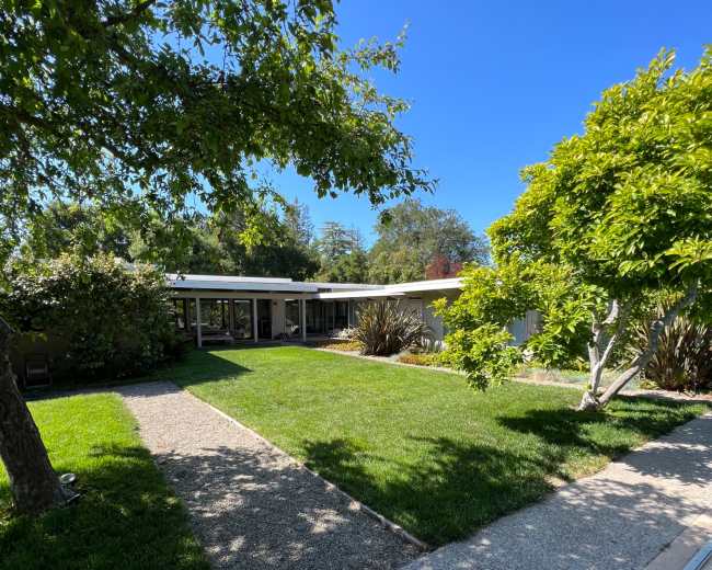 A modern house with a flat roof and large windows sits beside a swimming pool, surrounded by well-maintained grass and trees.