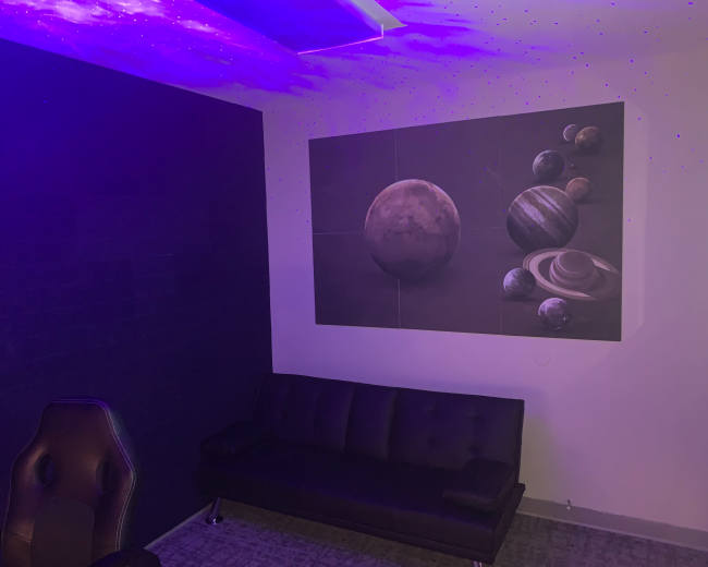 The image shows a room with a dark wall, a black couch, and a large mural of planets on the opposite wall, illuminated by purple lighting.