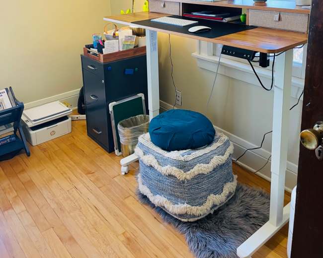 A standing desk with a monitor and lamp is positioned against a window in a room featuring a storage cabinet and decorative items on the walls.