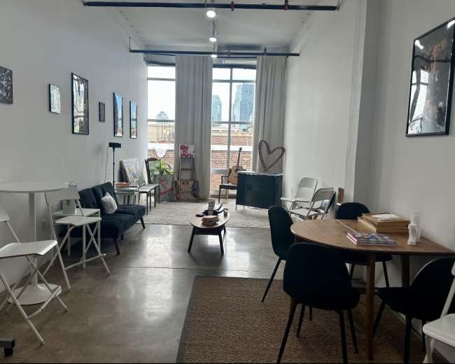 The image shows a spacious, well-lit room featuring a seating area with a black couch, a wooden table with chairs, and several pieces of wall art.