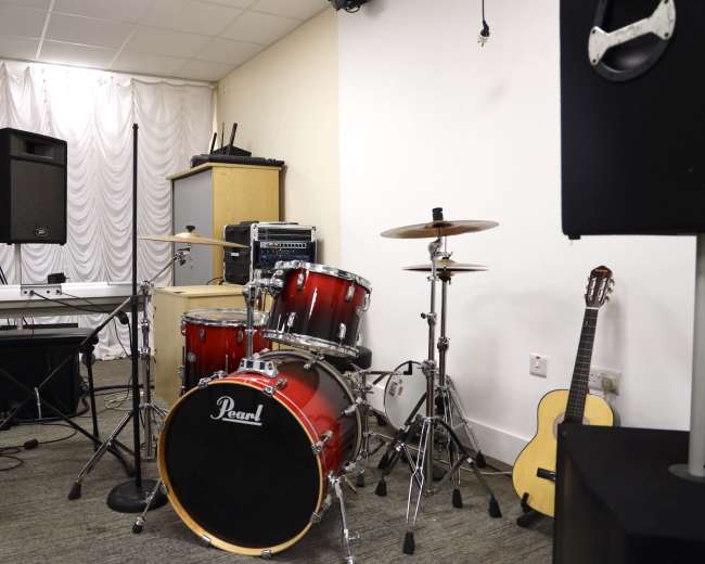 The image shows a small music setup featuring a drum kit, a keyboard, an amplifier, and an acoustic guitar against a wall in a room with white drapery.