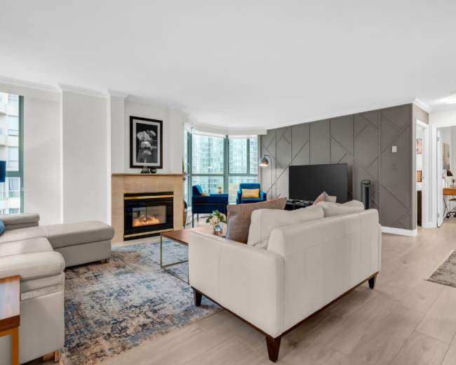 The image shows a modern living room with a fireplace, a large TV, and a mix of seating areas, including a light-colored sofa and an accent chair, set against large windows.