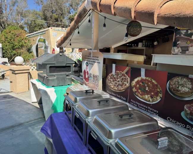 The scene features a pizza food stall with various pizza styles displayed on boards and warming trays arranged on a counter under a shaded area.