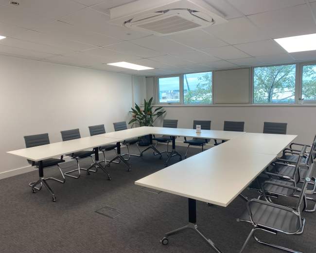 A conference room features a large, white rectangular table surrounded by multiple gray chairs, with windows showing greenery outside.