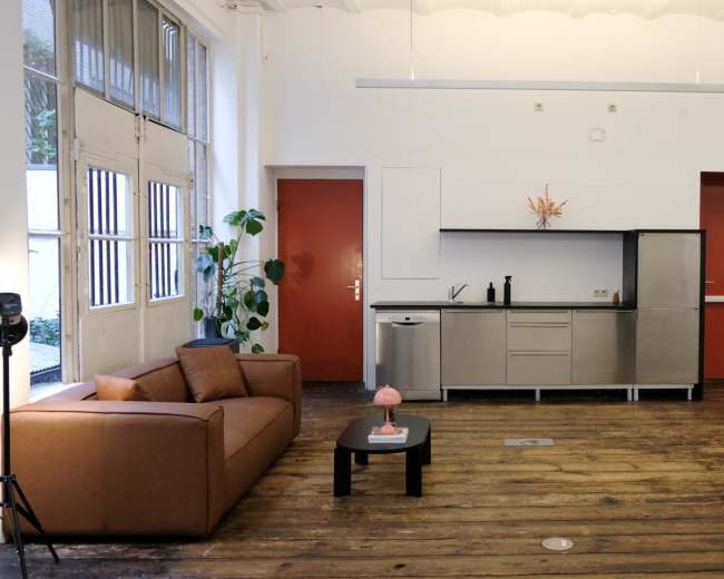 The image shows a spacious room featuring a brown sofa, a metal kitchen unit, and a tall wooden pillar, with red walls and large windows.