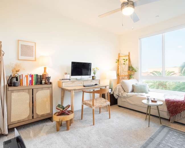 The image shows a cozy living room with a small desk, a sofa, a bookshelf, and a large window letting in natural light.