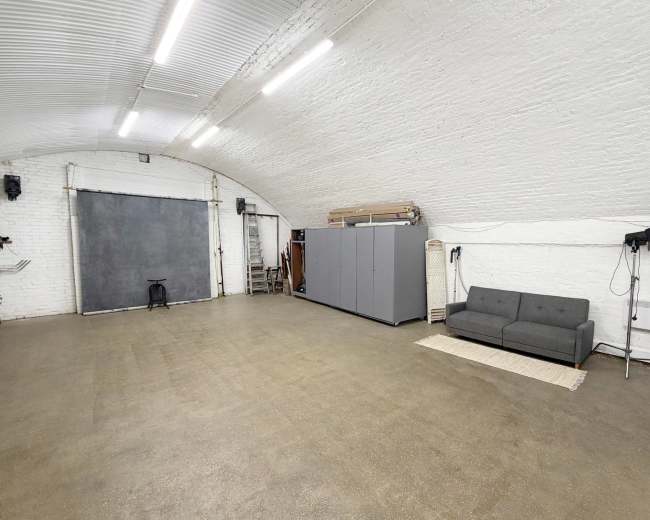 The image shows a spacious, empty room with white brick walls, a grey storage unit, a sofa, and a backdrop setup for photography.