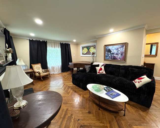The image shows a living room featuring a dark couch, a wooden coffee table, and large windows with decorative curtains.