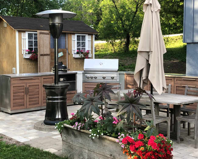 The image shows an outdoor kitchen area with a stainless steel grill, wooden cabinets, a patio table with chairs, a large umbrella, and a decorative planter filled with flowers.