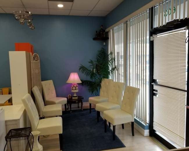 A waiting area with several upholstered chairs arranged around a small table, illuminated by a lamp, against a backdrop of a blue wall and large windows with blinds.
