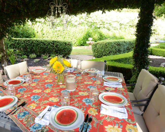 A neatly arranged dining table with plates of watermelon slices sits under a canopy of greenery, surrounded by a well-manicured garden.