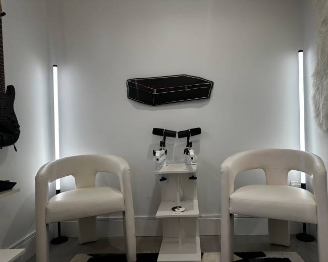 The image shows a small, modern room with two white chairs, a shelf with microscopes, and a wall displaying a guitar and an artwork resembling a suitcase.