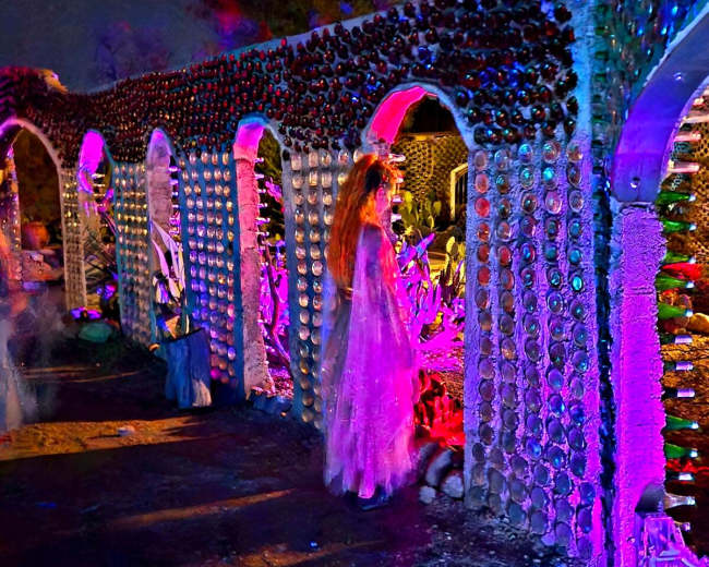 The scene depicts an illuminated outdoor walkway with arches made of bottles, surrounded by colorful lights and decorations.