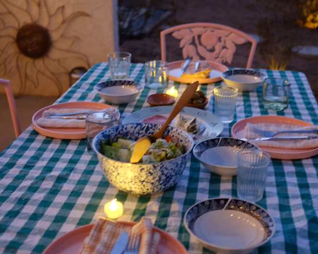 A dining table set with various dishware and a salad bowl is arranged outside near a vintage trailer at sunset.